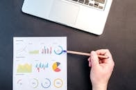 Hand holding pencil reviewing colorful data charts on desk with laptop.